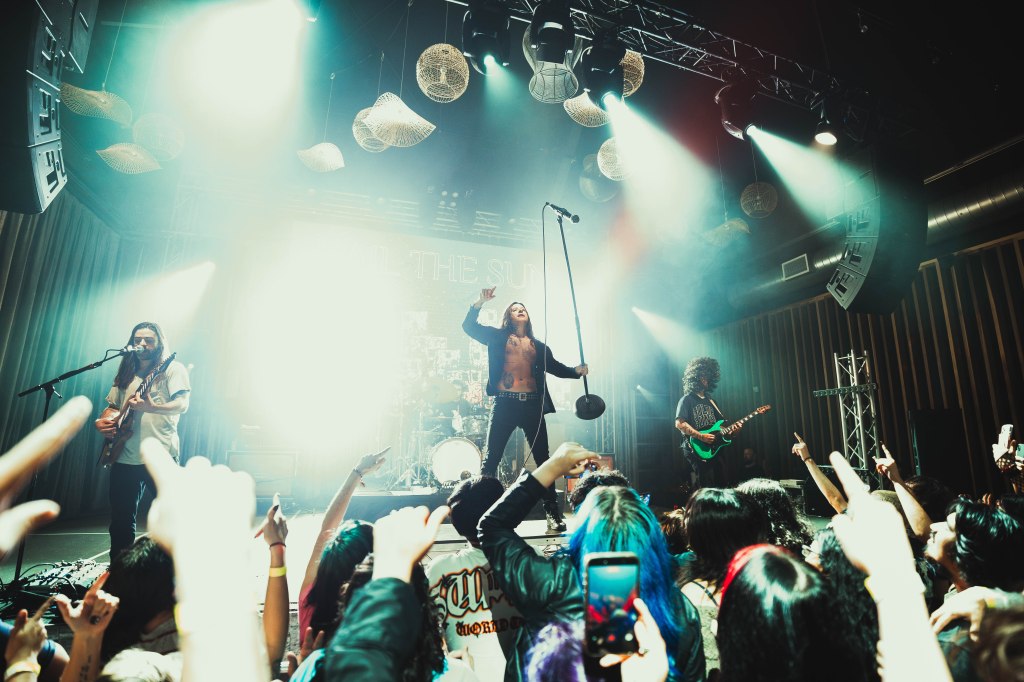 A rock band performing on stage with dramatic lighting, featuring a singer holding a microphone and two guitarists. The enthusiastic audience is raising their hands in excitement.