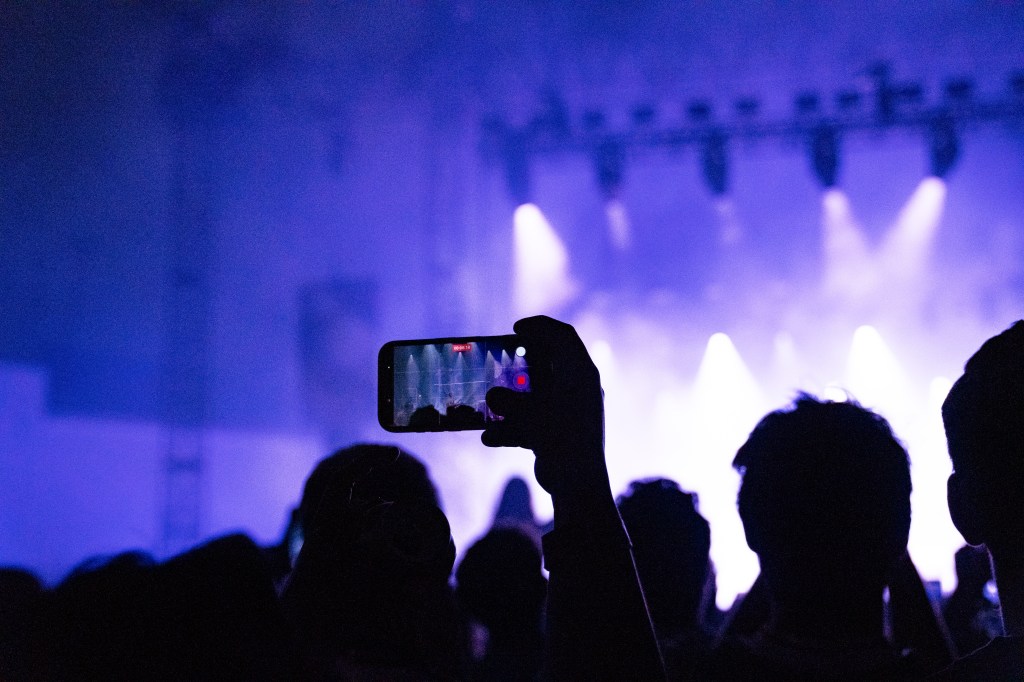 Audience at a concert with a silhouette of a person holding a smartphone to capture the performance, illuminated by purple stage lights.