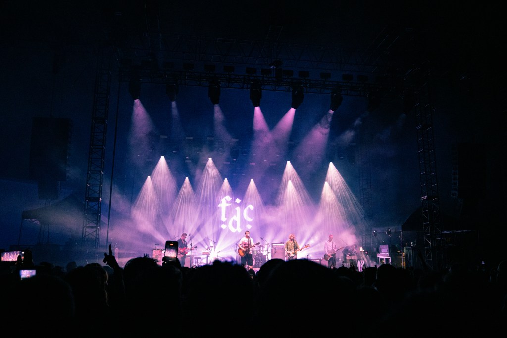 A live band performing on stage with vibrant purple lighting and a crowd in the foreground.