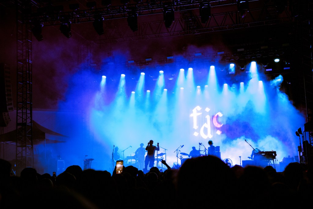 A concert scene with a silhouetted performer in front of illuminated stage lights and smoke, featuring the text 'fác' in the background.