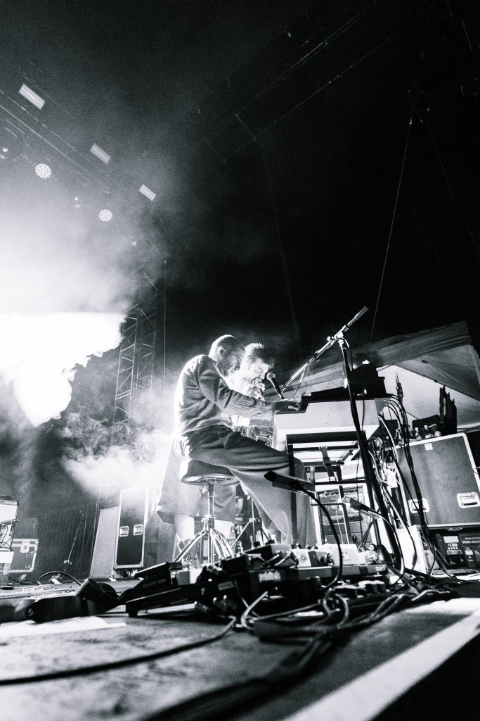 A musician performing at a concert, seated at a keyboard, with dramatic lighting and smoke effects in a black and white tone.