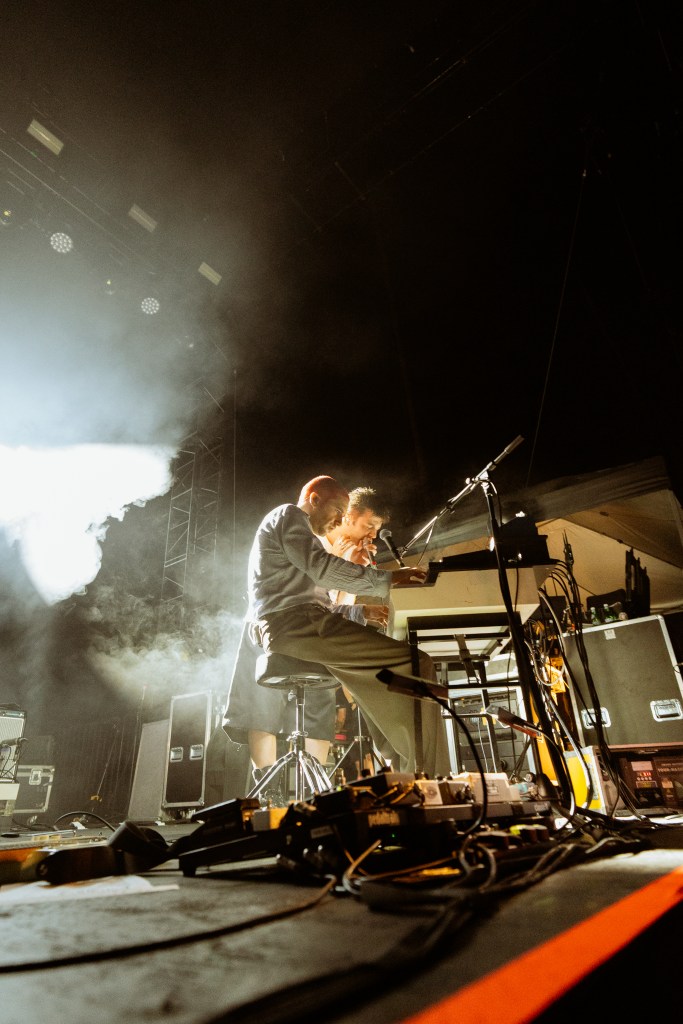 Two musicians performing on stage, one playing a keyboard and the other singing, with a dramatic lighting effect and smoke in the background.