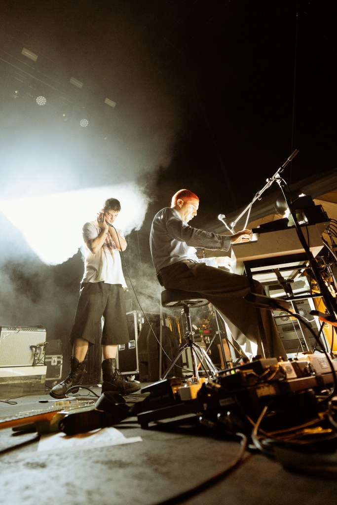 Two musicians performing on stage with instruments, surrounded by fog and stage lights.