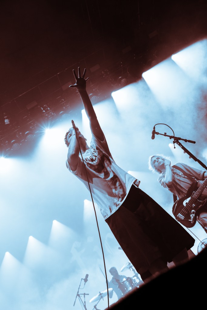 A singer on stage performing with one hand raised, wearing a graphic t-shirt and shorts, while a bassist plays nearby, against a backdrop of dramatic lighting.