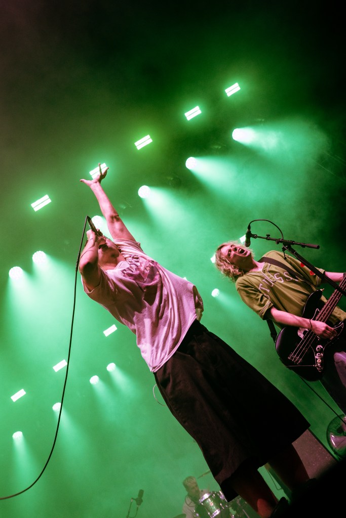 Two musicians performing on stage under green lighting, one holding a microphone and raising their arm enthusiastically, and the other playing a bass guitar.