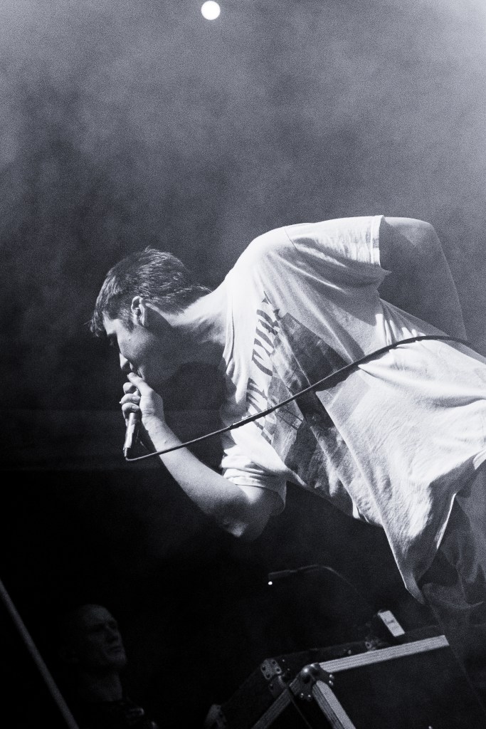 A performer singing into a microphone on stage, surrounded by fog and dramatic lighting. In the background, another person is partially visible.