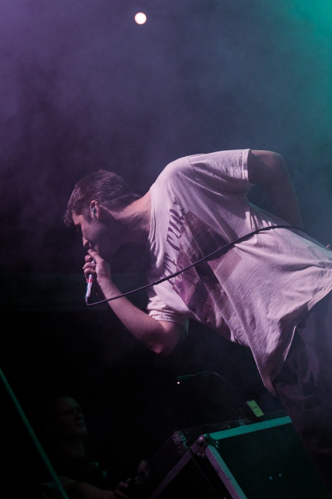 A musician performing on stage, holding a microphone and leaning forward, surrounded by colorful smoke and stage lighting.