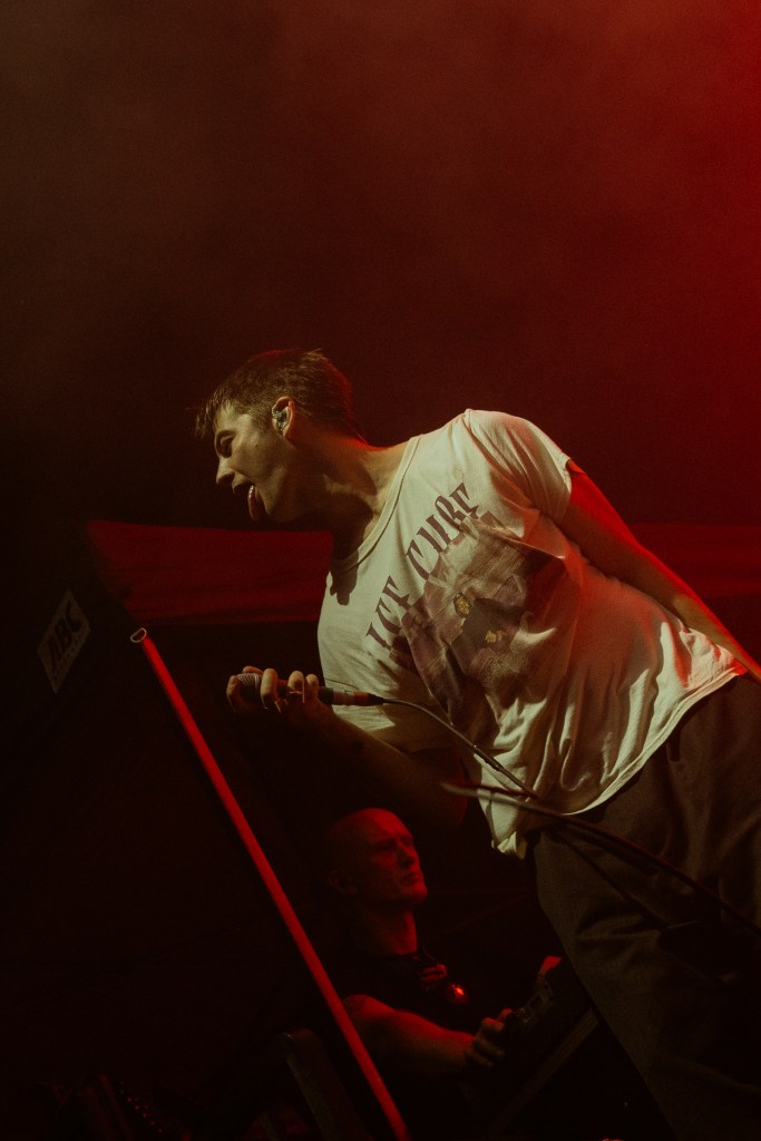 A male singer performing on stage, wearing a white t-shirt and jeans, with a dramatic expression and a microphone in hand. The background is illuminated with red lighting, creating an energetic atmosphere. A band member is visible in the background.