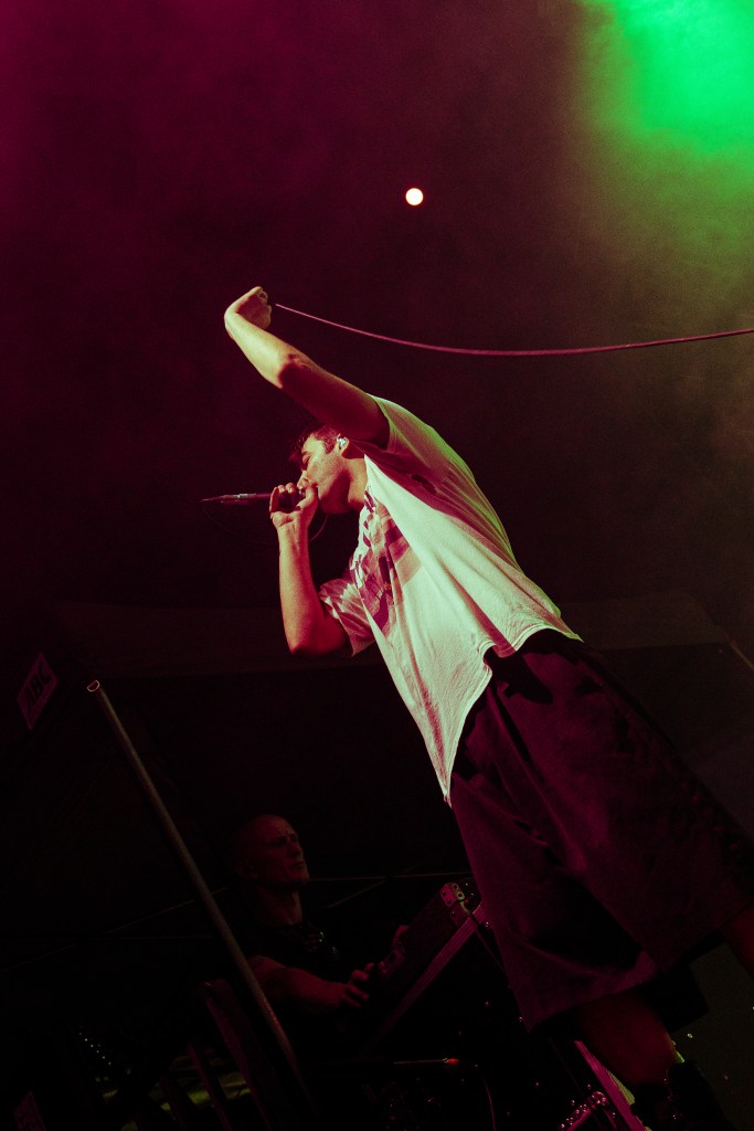 A performer sings into a microphone on stage, with dramatic lighting in purple and green hues. In the background, a sound technician can be seen managing equipment.