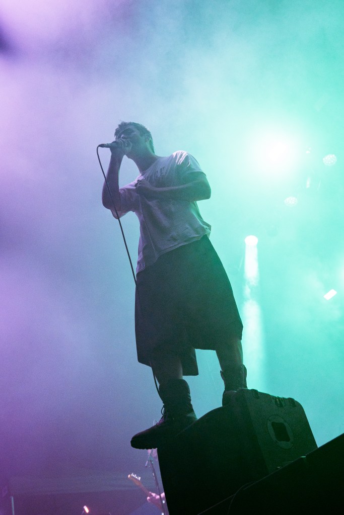 A singer performing on stage, standing on a speaker with a microphone in hand, surrounded by colorful lighting and smoke.