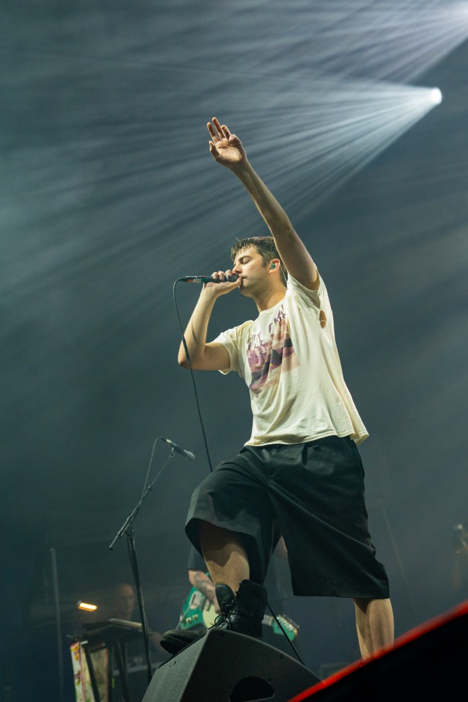 A male performer singing into a microphone with one hand raised, wearing a loose white t-shirt and black shorts, on stage during a concert with dramatic lighting.