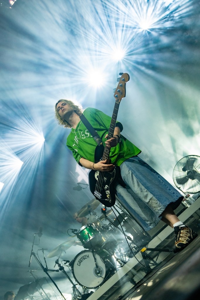A musician playing the bass guitar on stage, wearing a green shirt and oversized denim shorts, with dramatic lighting in the background.