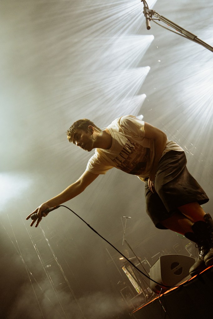 A male singer performing on stage with a microphone, wearing a t-shirt and shorts, surrounded by dramatic stage lighting.