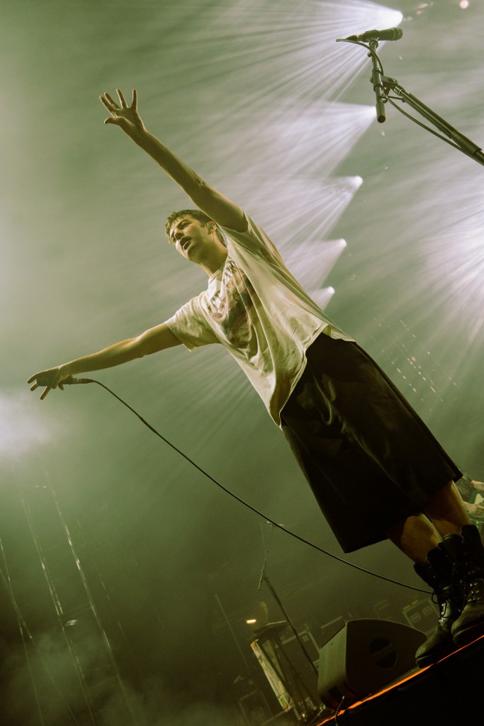 A male musician performing on stage with outstretched arms, wearing a loose white shirt and black shorts, against a backdrop of dramatic lighting.