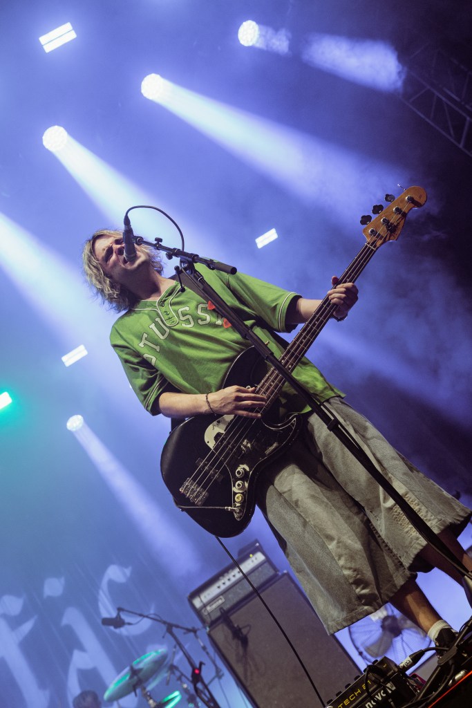A musician performing on stage with a bass guitar while singing into a microphone, dressed in a green t-shirt and denim shorts, with colorful stage lights in the background.