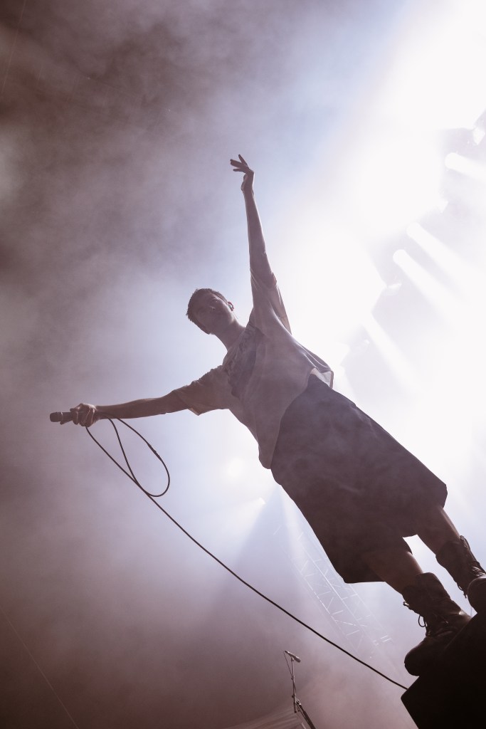 A silhouetted performer on stage with one arm raised, holding a microphone, surrounded by dramatic lighting and fog.