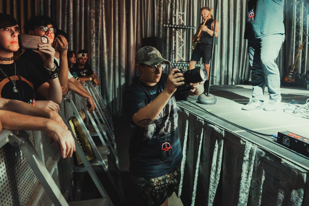 A photographer captures a moment at a live music event, surrounded by enthusiastic fans watching the performance. The stage is illuminated by lights, and musicians are visible in the background.