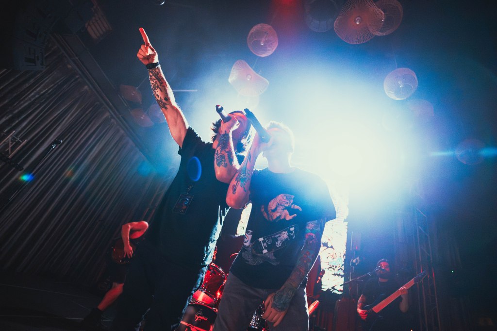 Two male vocalists performing on stage with microphones, surrounded by bright stage lights and band members in the background.