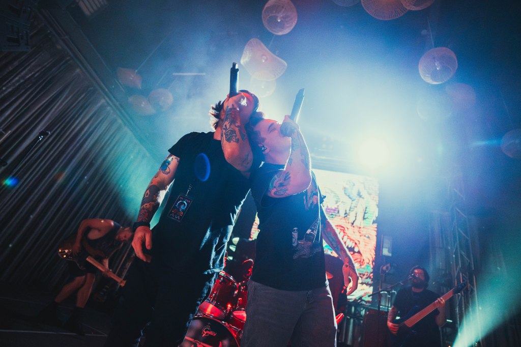 Two tattooed male vocalists performing on stage, microphones in hand, with a colorful light display and band members visible in the background.