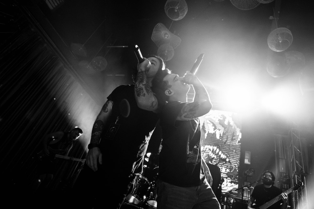 Two male performers singing into microphones on stage, illuminated by bright lights, with band members visible in the background.