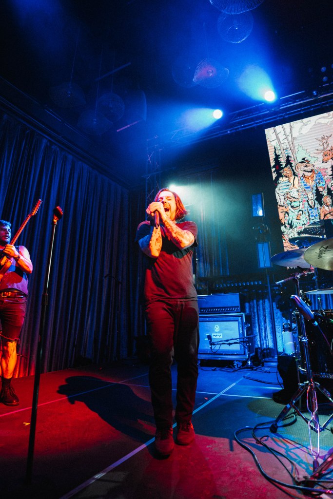 A lead singer passionately performing on stage with a microphone, surrounded by colorful stage lighting, while a guitarist is visible in the background.