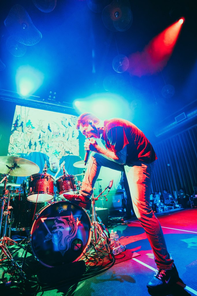 A male vocalist performing energetically on stage, with a drum set in the background and colorful stage lighting.