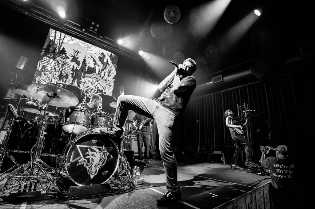 A musician passionately performing on stage with a microphone, surrounded by band members playing instruments. The background features a dramatic visual projection, and the image is in black and white.