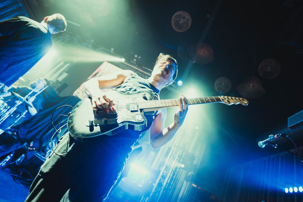 Musician playing electric guitar on stage with dramatic lighting, surrounded by band members.