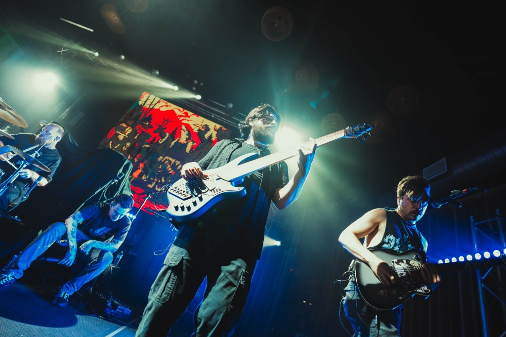 A band performing on stage with bright lights; a bassist playing a white bass guitar, a guitarist in a sleeveless shirt, and a drummer in the background.