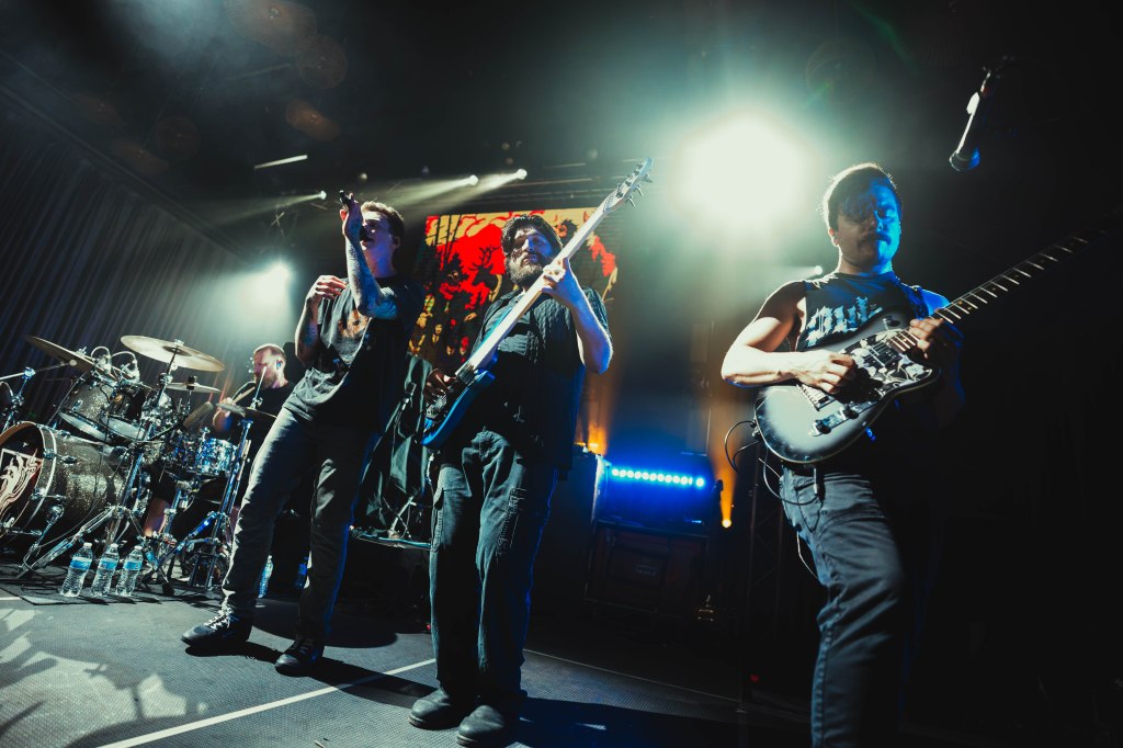A rock band performing live on stage with dramatic lighting, featuring a vocalist, guitarist, and drummer.
