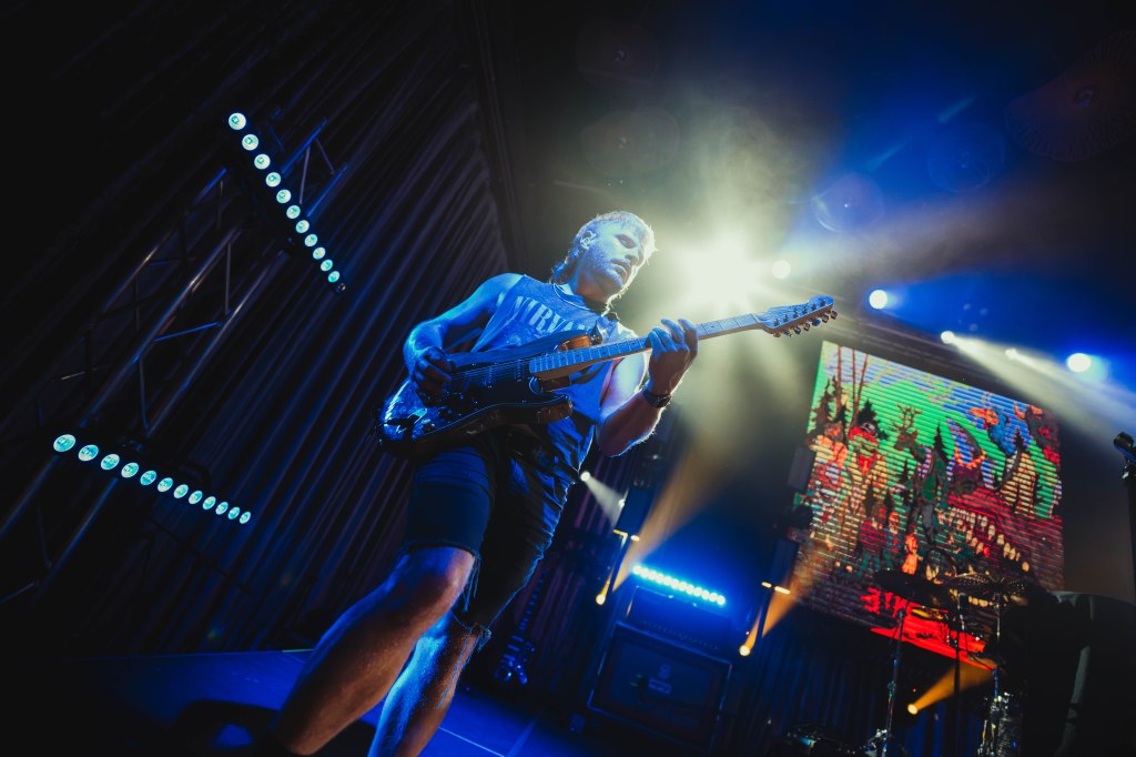 A guitarist performing on stage with dramatic lighting and colorful visuals in the background.