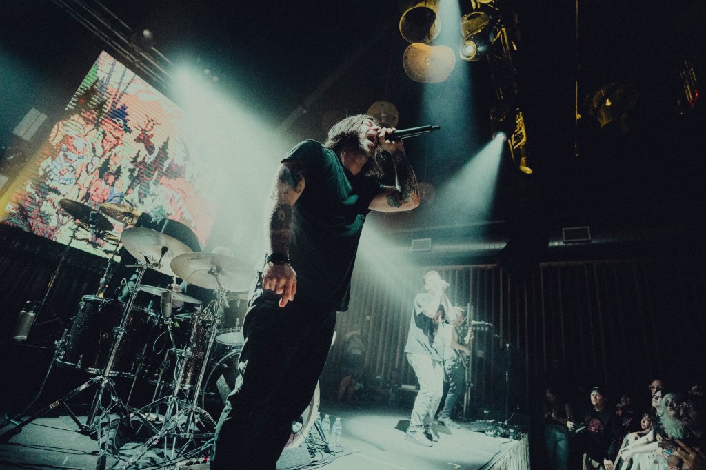 A live music performance with a vocalist singing into a microphone on stage, surrounded by band members, with a colorful backdrop and bright lights illuminating the scene.
