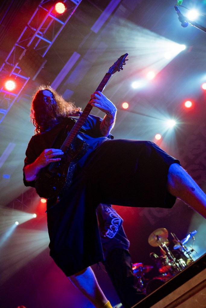 A guitarist performing on stage with dramatic lighting, playing a black electric guitar. The musician has long hair and tattoos, dressed in loose-fitting clothing.
