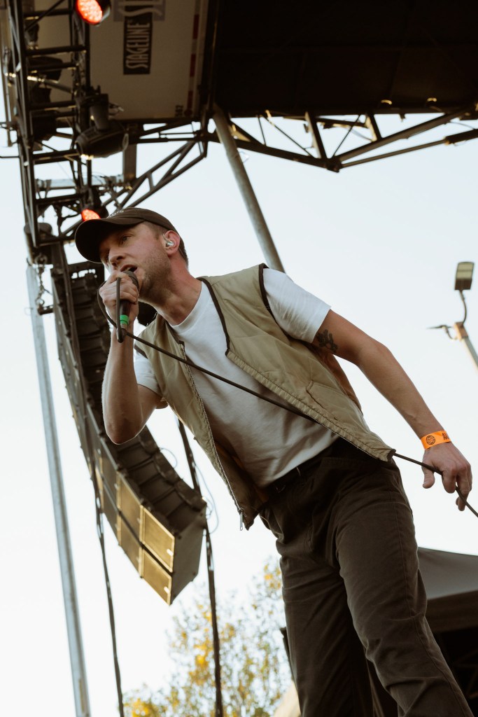 A male performer is singing into a microphone on stage, wearing a beige vest, white t-shirt, and a cap. The background features sound equipment and trees under a clear sky.