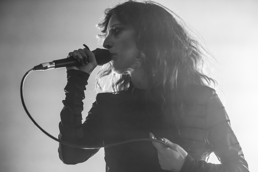 A singer performing on stage, holding a microphone and engaging with the audience, in a dramatic black and white setting.