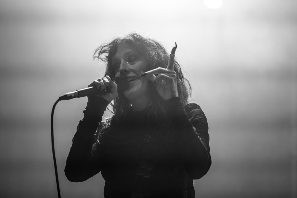 A performer singing into a microphone on stage, wearing a black outfit and gesturing with one hand, with a misty background in black and white.