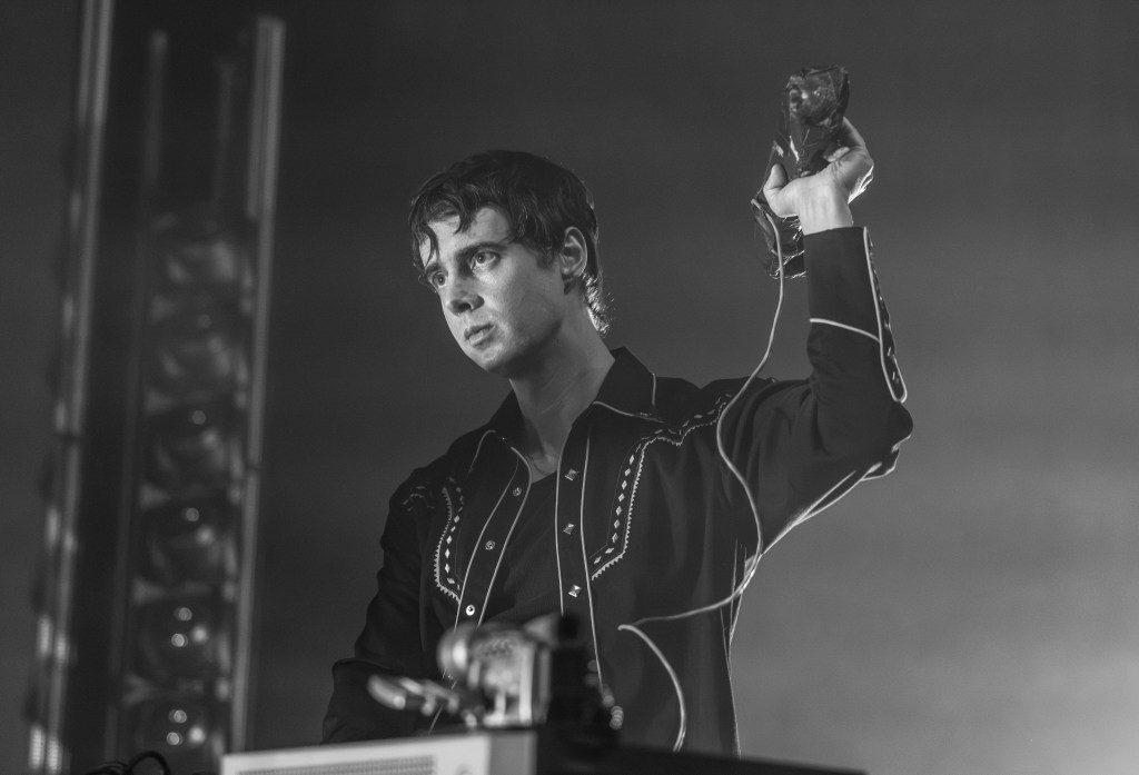 A musician performing on stage, wearing a black shirt with white embroidery, holding a microphone or instrument in one hand while looking intently at the audience.