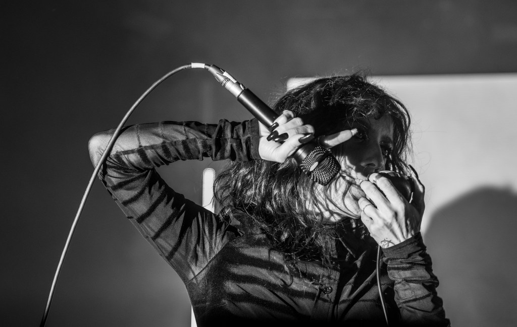 A performer passionately singing into a microphone with one hand while holding a hand to their face, dressed in a black long-sleeve shirt, captured in a dramatic monochrome style.