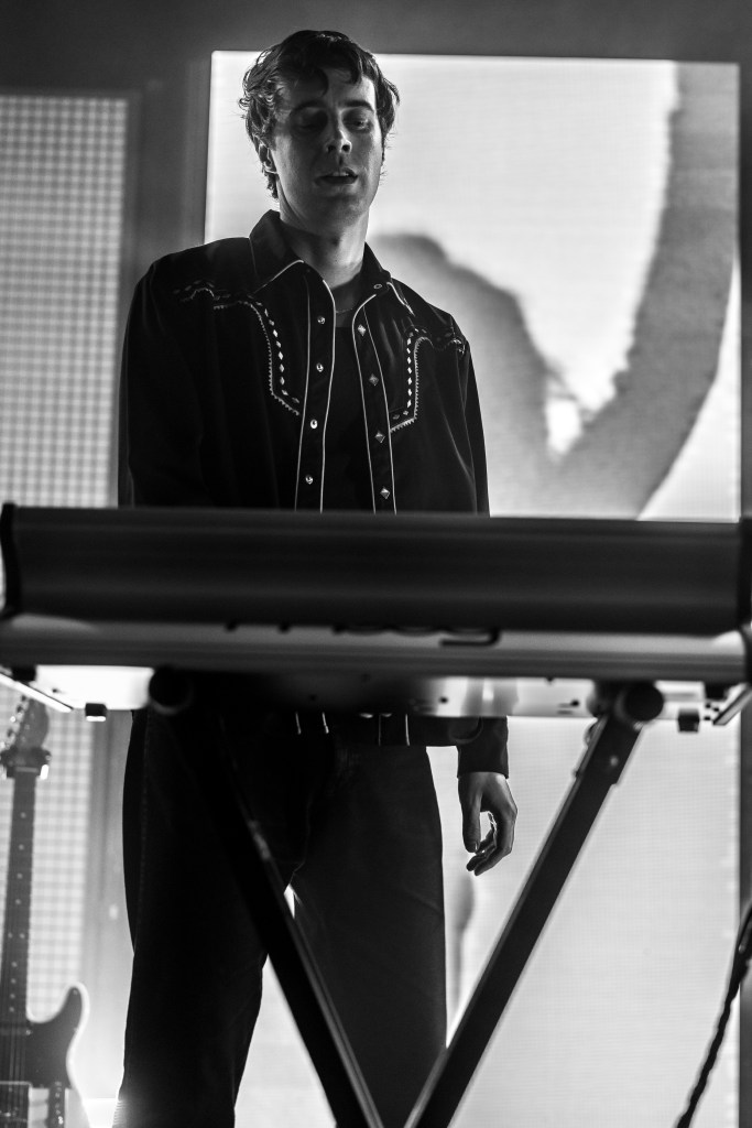 A musician performing on stage, playing a keyboard, wearing a black shirt with decorative stitching. The image is in black and white, highlighting the focused expression of the musician.