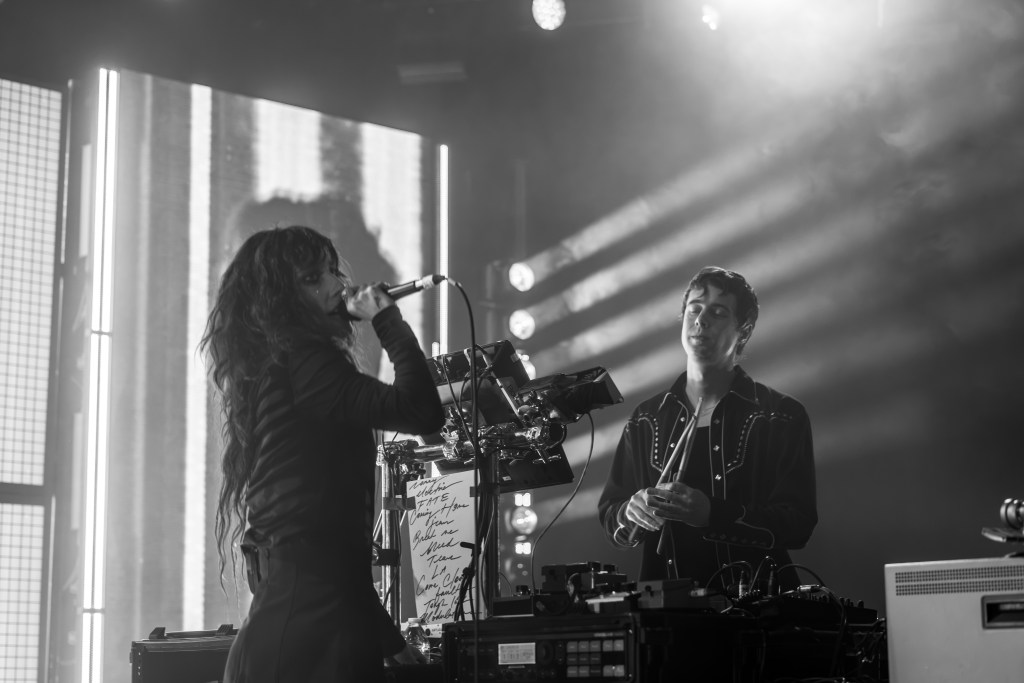 A musician performing on stage, holding a microphone, with long hair and wearing a black outfit. Another person is behind a music setup, looking at the performer while holding a drumstick. The scene is illuminated with dramatic lighting.