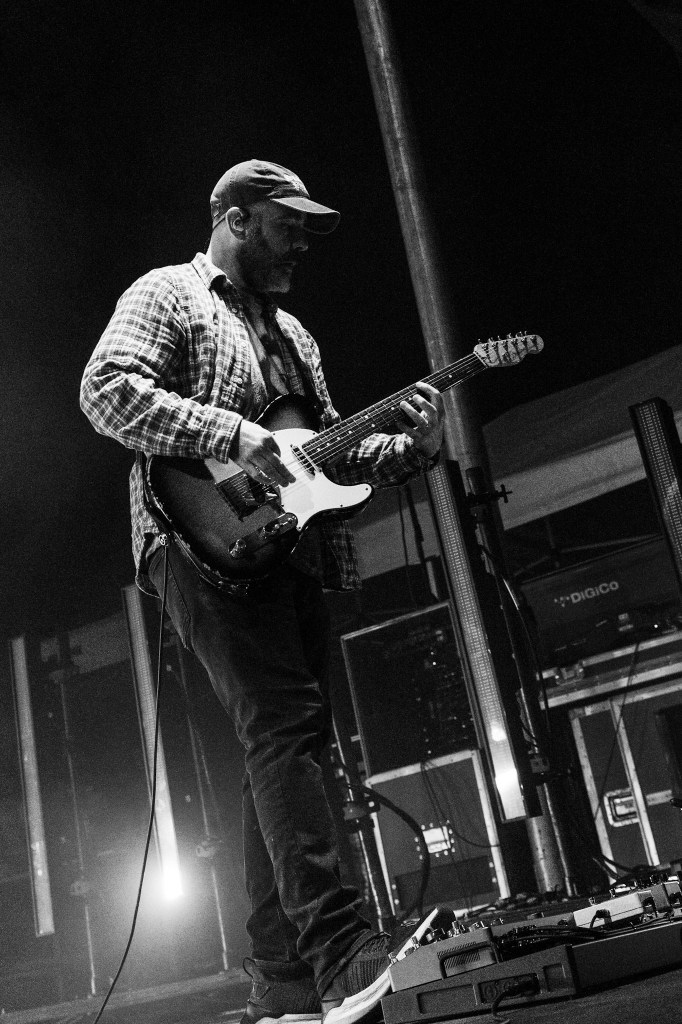A musician playing an electric guitar on stage, wearing a checkered shirt and a cap, surrounded by concert lighting and equipment.
