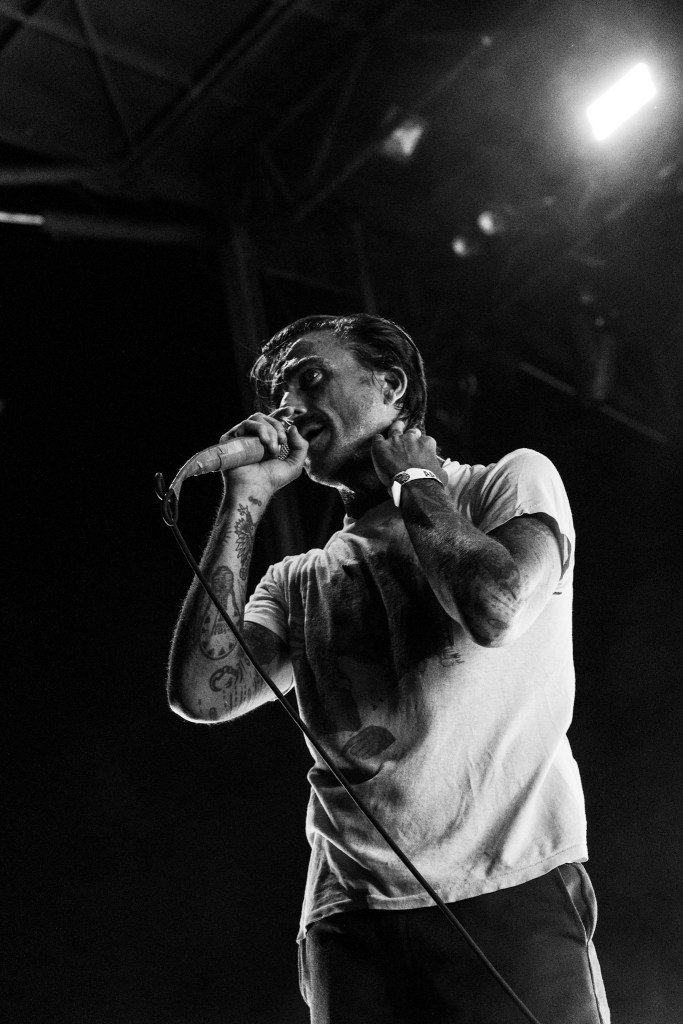A musician passionately singing into a microphone on stage, dressed in a casual t-shirt, with visible tattoos and intense facial expression, illuminated by stage lights.