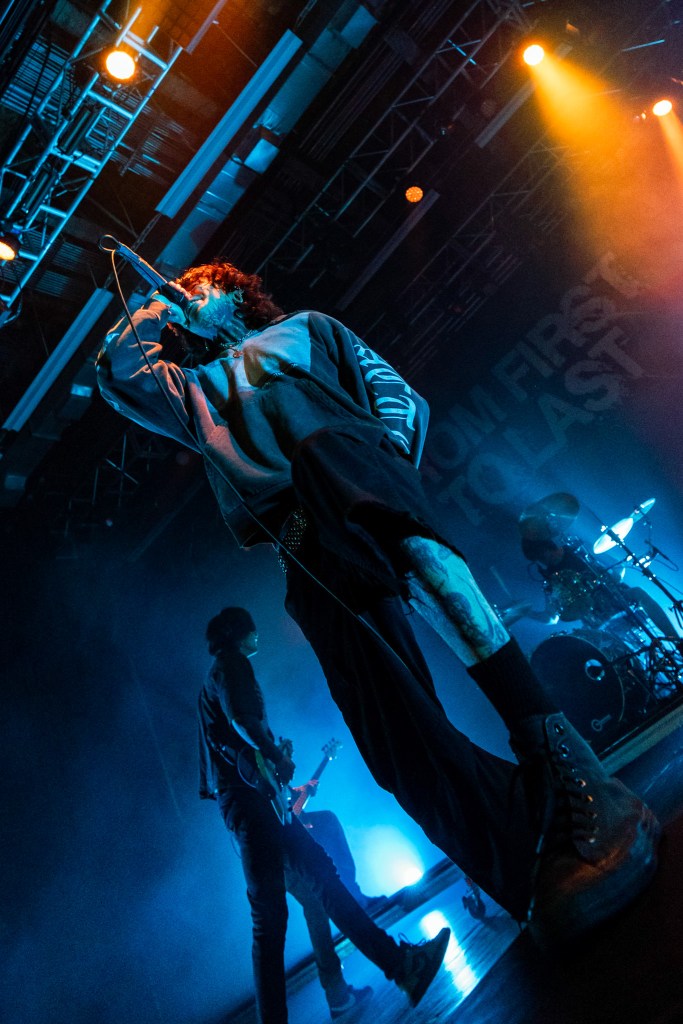 A musician performing on stage with a microphone, illuminated by dramatic lighting, surrounded by band members playing instruments.