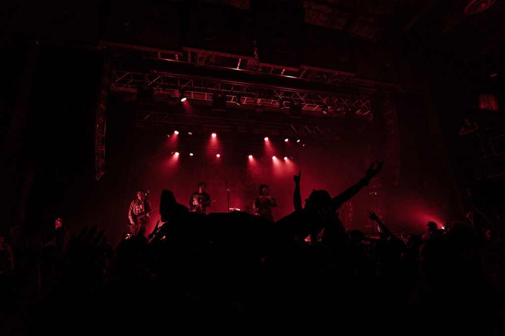 A silhouetted view of musicians performing on stage with red lighting, while an audience member crowdsurfs above the crowd.