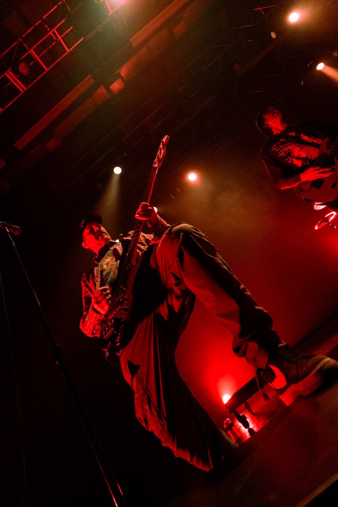 A musician performing on stage, focused on playing the bass guitar, with dramatic red lighting and another guitarist in the background.