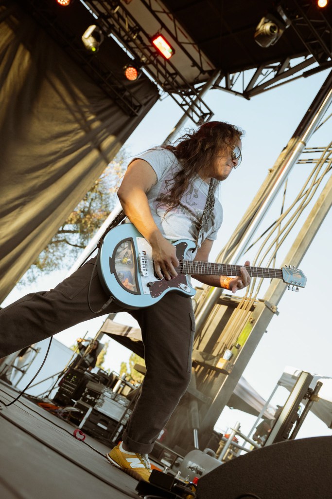A musician performing on stage with an electric guitar, wearing a gray t-shirt and black pants, with long hair and sunglasses.