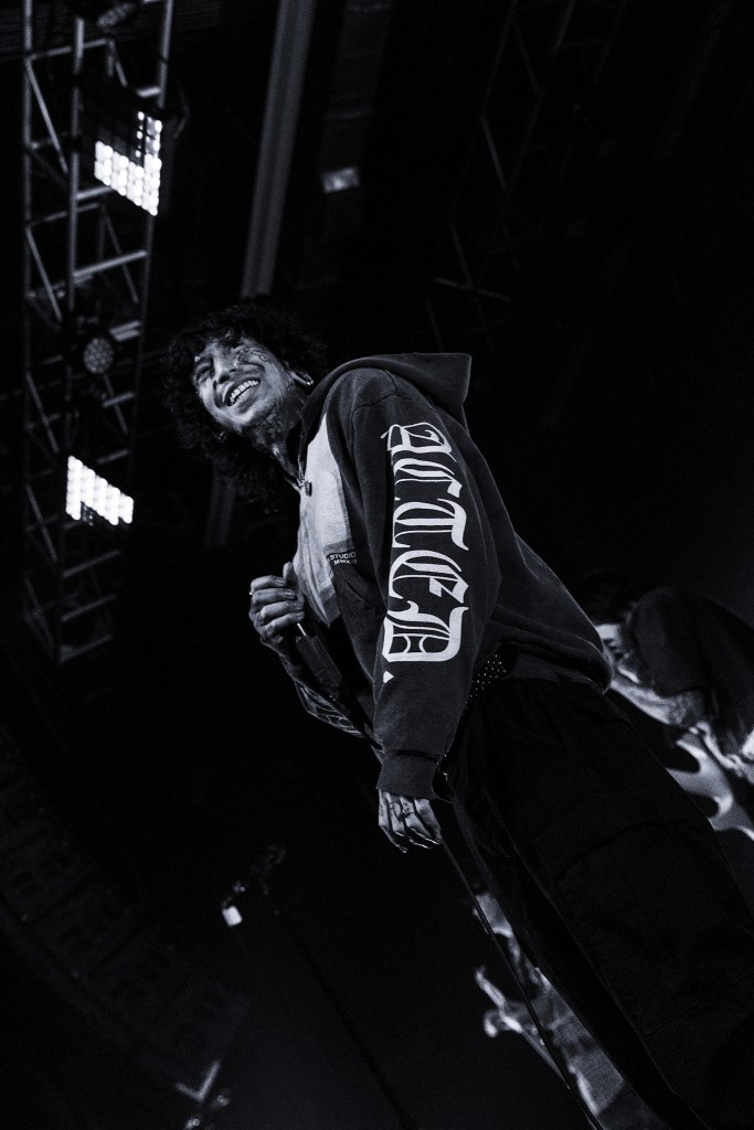 A performer on stage smiling widely while wearing a black hoodie with a graphic design, captured in black and white.