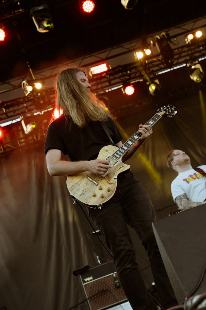 A musician with long hair playing an electric guitar on stage, with bandmates and stage lights in the background.