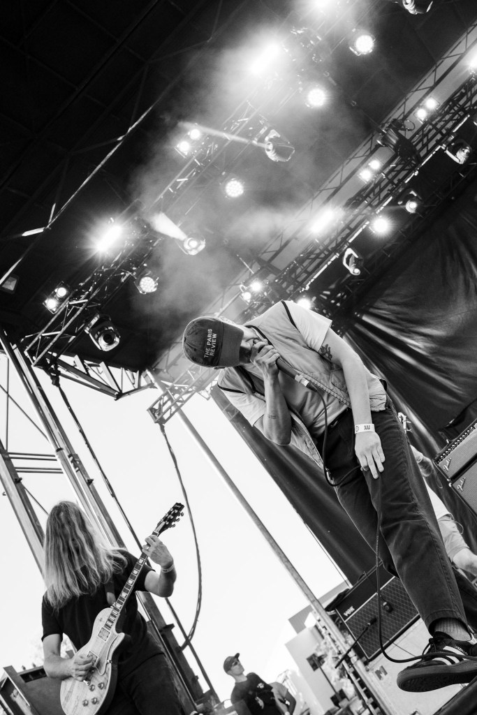 A musician performing on stage, passionately singing into a microphone while another band member plays electric guitar in the background. The lighting creates a dramatic atmosphere with smoke effects.