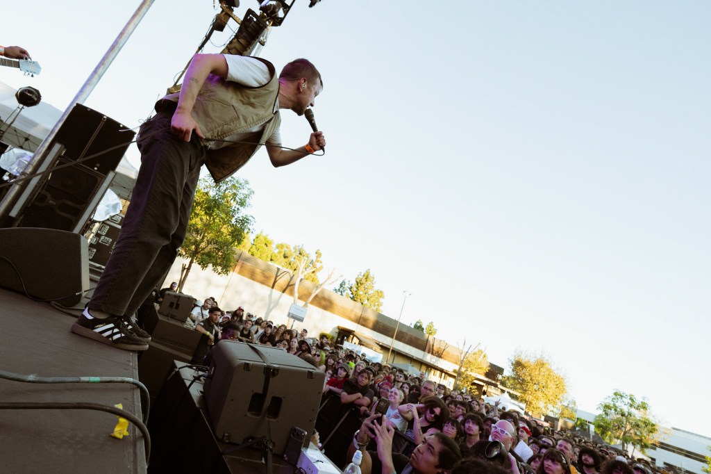 A performer energetically singing into a microphone on stage, with a crowd of enthusiastic fans in front. The scene is set outdoors with clear skies and trees in the background.